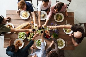 Friends sitting at a wooden dinner table all raising their glasses at the same time to make a toast, with plates of food and bread on the table.