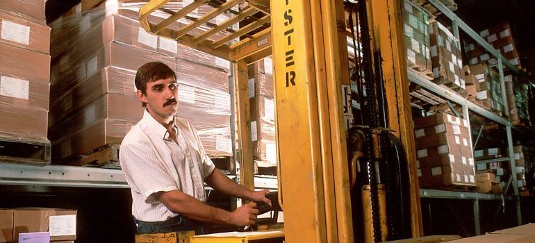 A man operating a forklift between pallets of boxes in warehouse storage