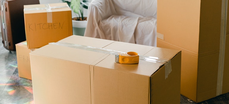 Taped moving boxes, one labeled kitchen, with a tape dispenser and a sheet-covered armchair