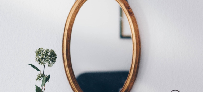Oval mirror in a gold wooden frame hanging on a white wall next to a green plant
