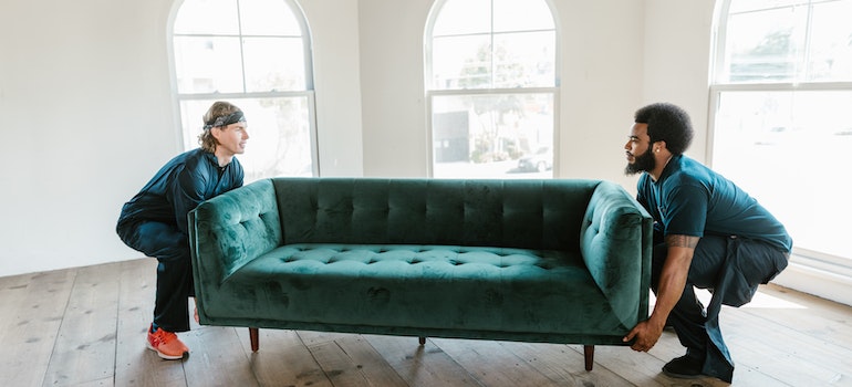 Two movers squatting to lift a green velvet sofa from both ends in an empty room