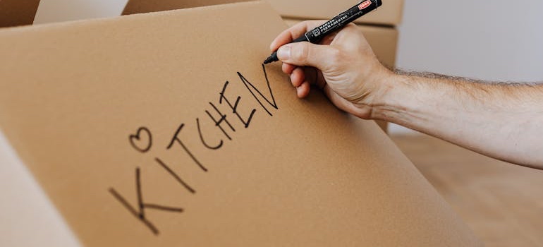 A person writing the word kitchen on a moving box with a marker
